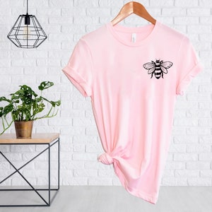 May include: A light pink short-sleeved t-shirt featuring a black graphic of a bee on the upper left chest area. The shirt has a crew neckline and is displayed on a wooden hanger with a knot tied at the bottom hem to create a draped effect.