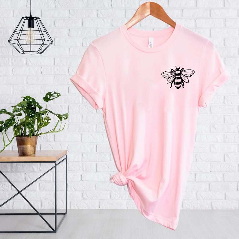 May include: A light pink short-sleeved t-shirt featuring a black graphic of a bee on the upper left chest area. The shirt has a crew neckline and is displayed on a wooden hanger with a knot tied at the bottom hem to create a draped effect.