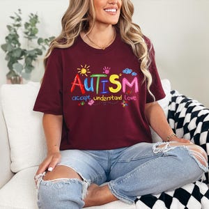 May include: A maroon t-shirt with the word "AUTISM" in colorful embroidered letters. Below the word are the words "accept understand love". The shirt has a crew neck and short sleeves.