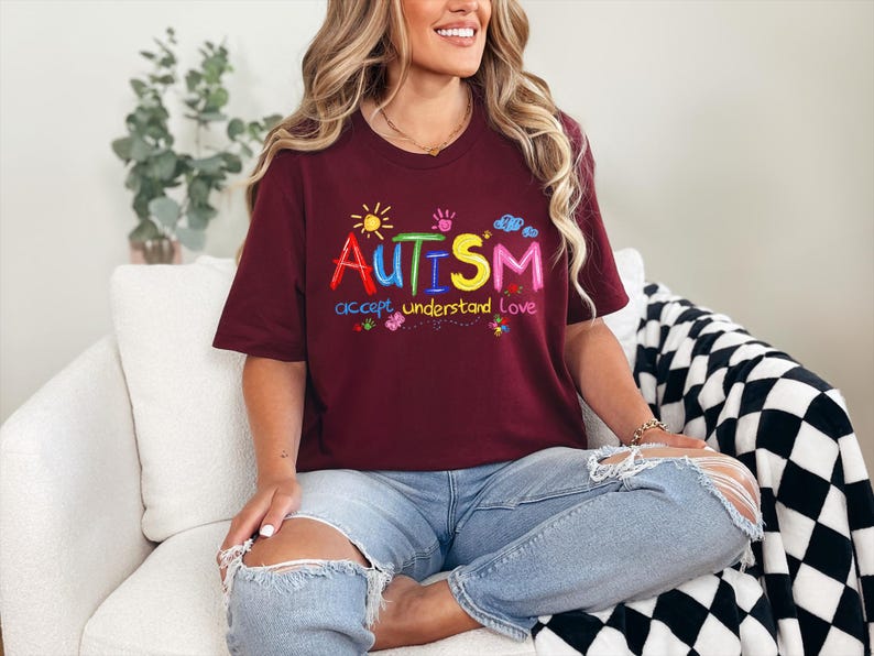 May include: A maroon t-shirt with the word "AUTISM" in colorful embroidered letters. Below the word are the words "accept understand love". The shirt has a crew neck and short sleeves.