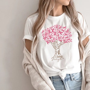 May include: A white short-sleeved t-shirt featuring a graphic of a tree where the leaves are composed of pink awareness ribbons. The trunk of the tree contains the words faith, hope, cure, grace, and future strength in a brown script font.