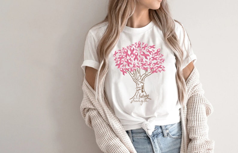 May include: A white short-sleeved t-shirt featuring a graphic of a tree where the leaves are composed of pink awareness ribbons. The trunk of the tree contains the words faith, hope, cure, grace, and future strength in a brown script font.
