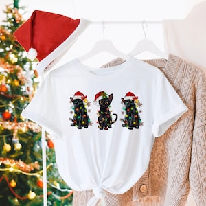Pode incluir: Camiseta branca com um design de Natal com tr&ecirc;s gatos pretos usando chap&eacute;us de Papai Noel e envoltos em luzes de Natal.