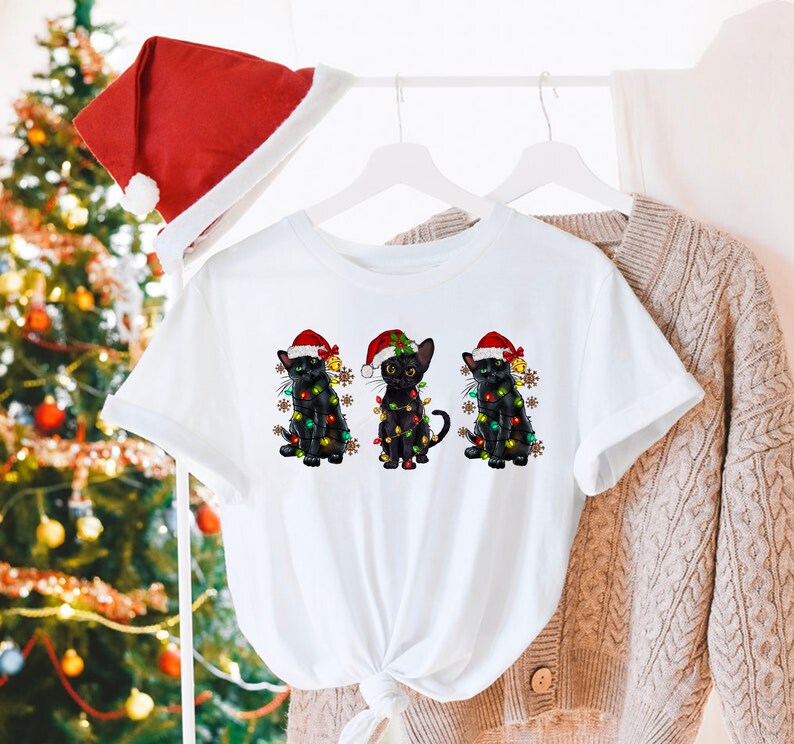 Pode incluir: Camiseta branca com um design de Natal com tr&ecirc;s gatos pretos usando chap&eacute;us de Papai Noel e envoltos em luzes de Natal.