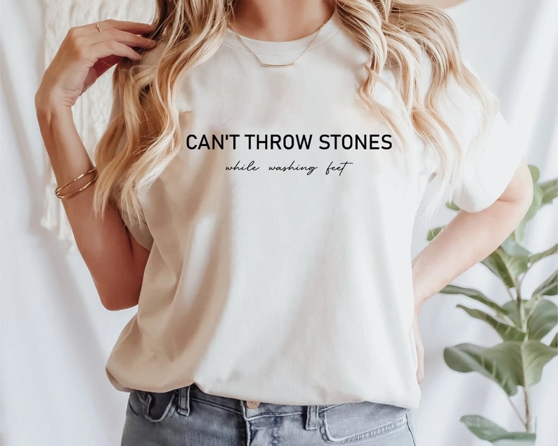 Puede incluir: Una camiseta blanca con el texto "CAN'T THROW STONES while washing feet" impreso en ella.