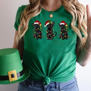 Pode incluir: Camiseta verde com tr&ecirc;s gatos pretos usando chap&eacute;us de Papai Noel e luzes de Natal. Os gatos est&atilde;o dispostos em fila, com o gato do meio olhando para a frente e os outros dois gatos olhando para os lados.