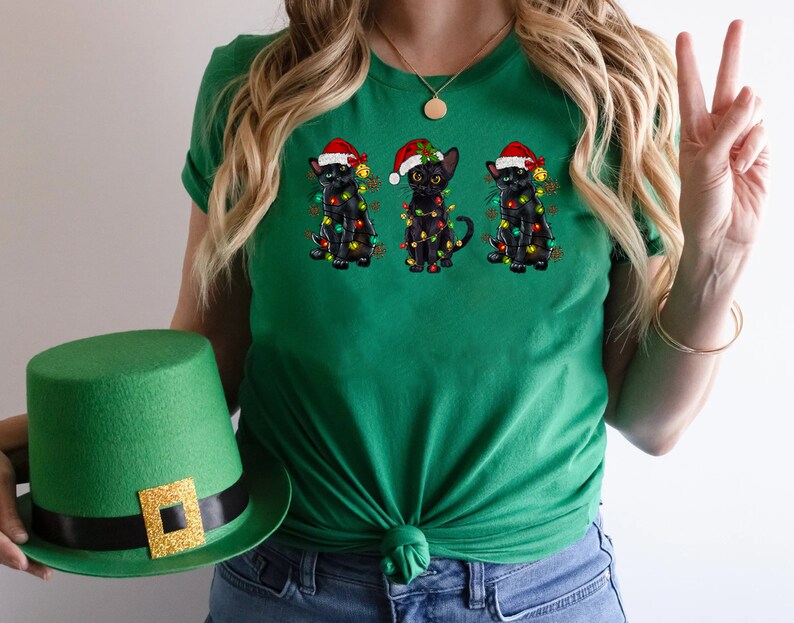 Pode incluir: Camiseta verde com tr&ecirc;s gatos pretos usando chap&eacute;us de Papai Noel e luzes de Natal. Os gatos est&atilde;o dispostos em fila, com o gato do meio olhando para a frente e os outros dois gatos olhando para os lados.