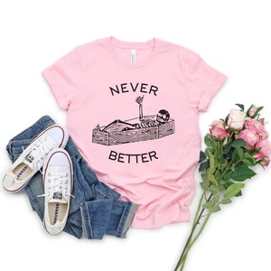 May include: Pink t-shirt with a black graphic of a skeleton lying in a coffin with the text "NEVER BETTER" printed on the shirt.
