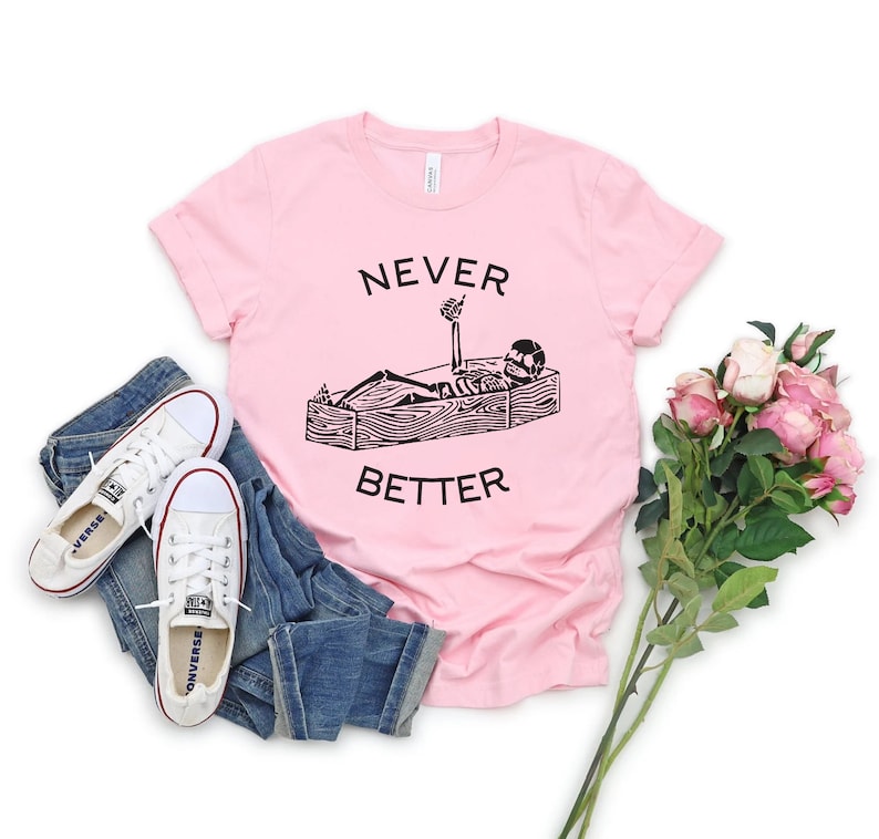 May include: Pink t-shirt with a black graphic of a skeleton lying in a coffin with the text "NEVER BETTER" printed on the shirt.