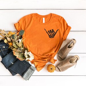 Op de afbeelding: Oranje T-shirt met een zwarte skelet hand die een shaka-teken maakt. Het shirt ligt op een wit houten oppervlak met een blauwe spijkerbroek, een kleine pompoen en herfstkleurige bloemen.
