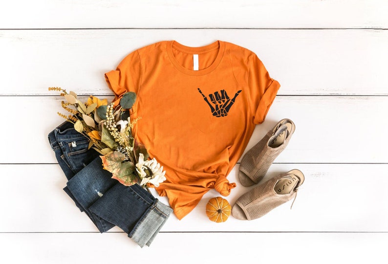 Op de afbeelding: Oranje T-shirt met een zwarte skelet hand die een shaka-teken maakt. Het shirt ligt op een wit houten oppervlak met een blauwe spijkerbroek, een kleine pompoen en herfstkleurige bloemen.