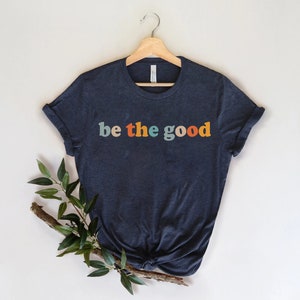 May include: Navy blue t-shirt with the text "be the good" in colorful retro font. The shirt is displayed on a wooden hanger against a neutral background, with a sprig of greenery adding a natural touch. A casual and inspirational apparel item.