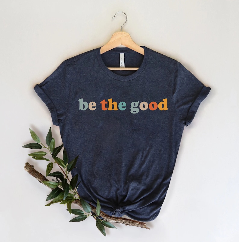 May include: Navy blue t-shirt with the text "be the good" in colorful retro font. The shirt is displayed on a wooden hanger against a neutral background, with a sprig of greenery adding a natural touch. A casual and inspirational apparel item.