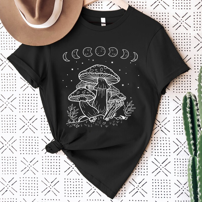 May include: Black t-shirt with a white outline of mushrooms and a crescent moon phase design. The shirt has short sleeves and a crew neck.