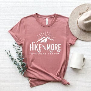 May include: A dusty rose t-shirt with the words "Hike More Worry Less" in white, featuring a mountain and paw print design. A beige hat and white mug are next to the shirt. A sprig of green leaves is also present.