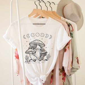May include: White t-shirt with a black and white illustration of mushrooms and a crescent moon phase design. The shirt is on a hanger.