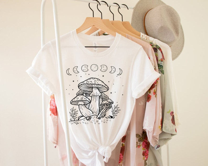 May include: White t-shirt with a black and white illustration of mushrooms and a crescent moon phase design. The shirt is on a hanger.