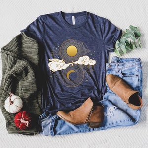May include: A navy blue t-shirt with a gold sun and moon design. The design features clouds and stars.