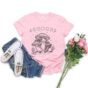 May include: A pink t-shirt with a black and white illustration of mushrooms and a crescent moon phase design. The shirt has short sleeves and a crew neck.
