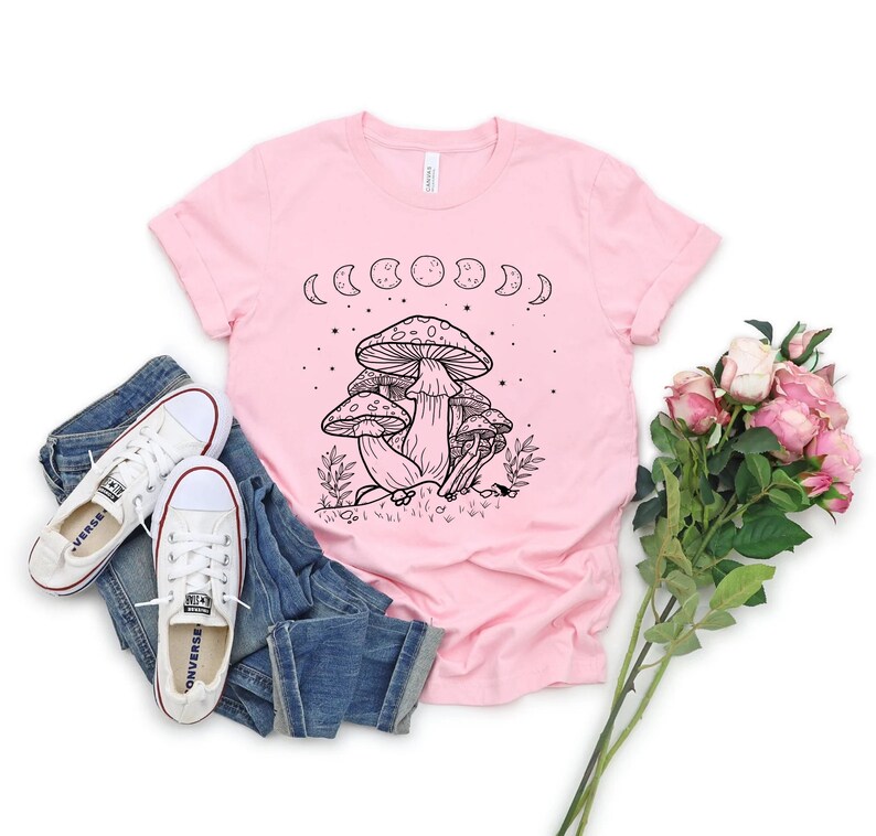 May include: A pink t-shirt with a black and white illustration of mushrooms and a crescent moon phase design. The shirt has short sleeves and a crew neck.