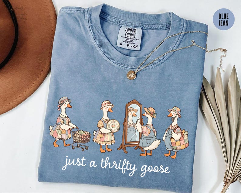 May include: Blue jean Comfort Colors t-shirt with a charming illustration of geese in vintage clothing. The geese are shown with shopping carts, mirrors, and baskets. The text "just a thrifty goose" is printed on the shirt.