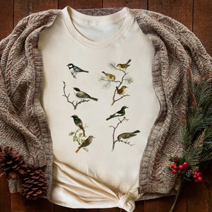 May include: Cream-colored t-shirt featuring detailed illustrations of various birds perched on branches. The shirt is paired with a brown cable-knit cardigan and decorative pinecones and greenery, creating a cozy, nature-inspired aesthetic.