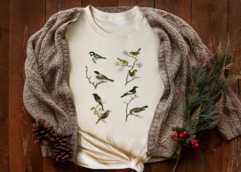 May include: Cream-colored t-shirt featuring detailed illustrations of various birds perched on branches. The shirt is paired with a brown cable-knit cardigan and decorative pinecones and greenery, creating a cozy, nature-inspired aesthetic.