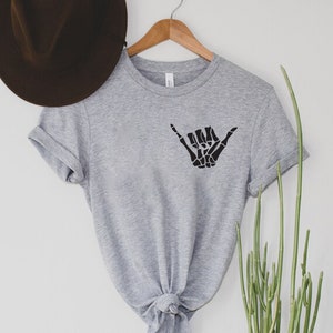 Op de afbeelding: Grijs T-shirt met korte mouwen met een zwarte skelethand die een shaka-teken maakt.