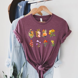 Może przedstawiać: Bordowy t-shirt z nadrukiem kwiatowym przedstawiającym dziewięć r&oacute;żnych kwiat&oacute;w w odcieniach pomarańczowego, ż&oacute;łtego, r&oacute;żowego i fioletowego. Kwiaty są ułożone w rzędzie na przodzie koszulki.