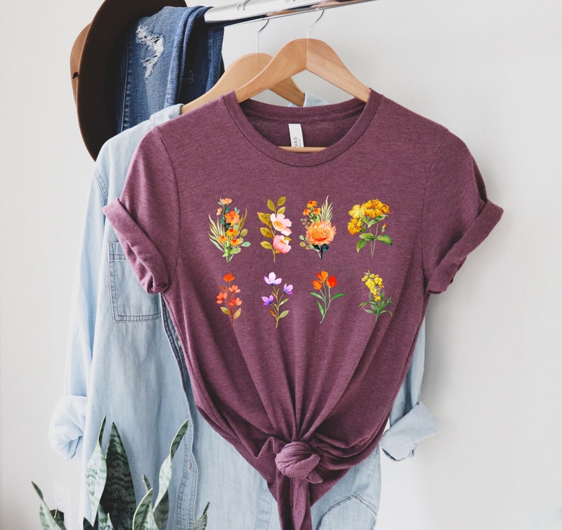 Może przedstawiać: Bordowy t-shirt z nadrukiem kwiatowym przedstawiającym dziewięć r&oacute;żnych kwiat&oacute;w w odcieniach pomarańczowego, ż&oacute;łtego, r&oacute;żowego i fioletowego. Kwiaty są ułożone w rzędzie na przodzie koszulki.