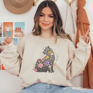 May include: Beige sweatshirt featuring a floral cat design. The cat is depicted in a seated position, with a colorful pattern of flowers and leaves. The sweatshirt has long sleeves and a crew neck.
