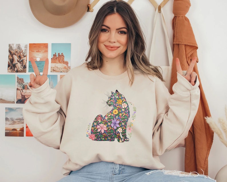 May include: Beige sweatshirt featuring a floral cat design. The cat is depicted in a seated position, with a colorful pattern of flowers and leaves. The sweatshirt has long sleeves and a crew neck.