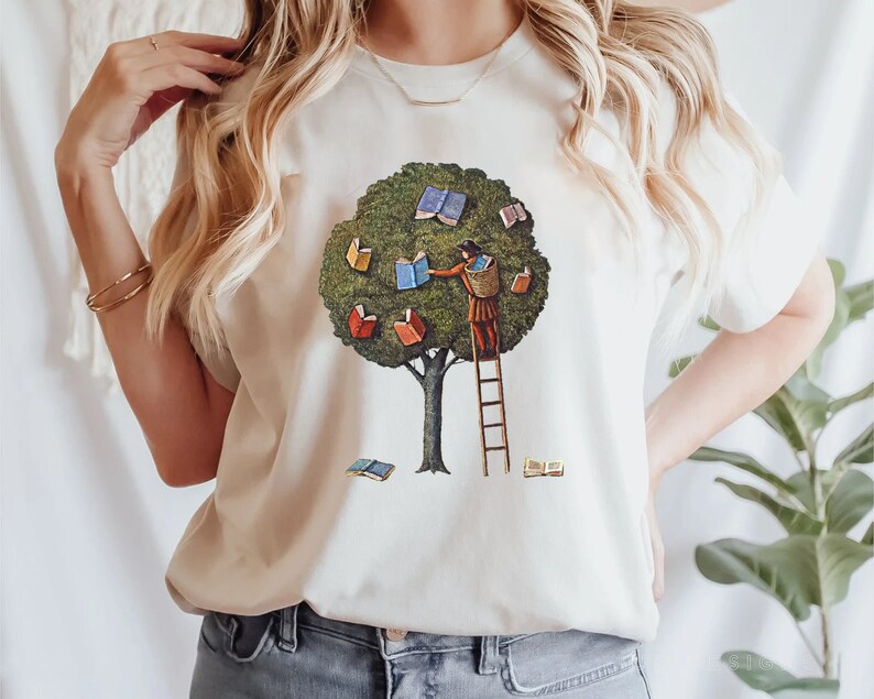 Pu&ograve; includere: Una maglietta bianca con una grafica di una persona che sale su una scala per raggiungere un libro in un albero. L'albero &egrave; pieno di libri con copertine colorate.