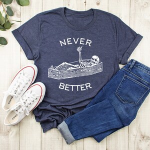 May include: A navy blue t-shirt with a white graphic of a skeleton lying in a coffin with the text "NEVER BETTER" printed on the shirt.