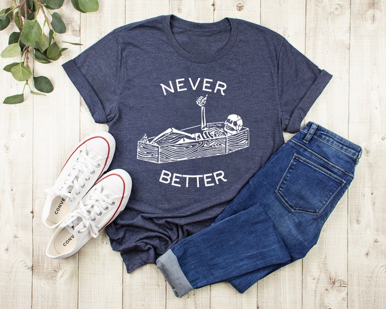 May include: A navy blue t-shirt with a white graphic of a skeleton lying in a coffin with the text "NEVER BETTER" printed on the shirt.
