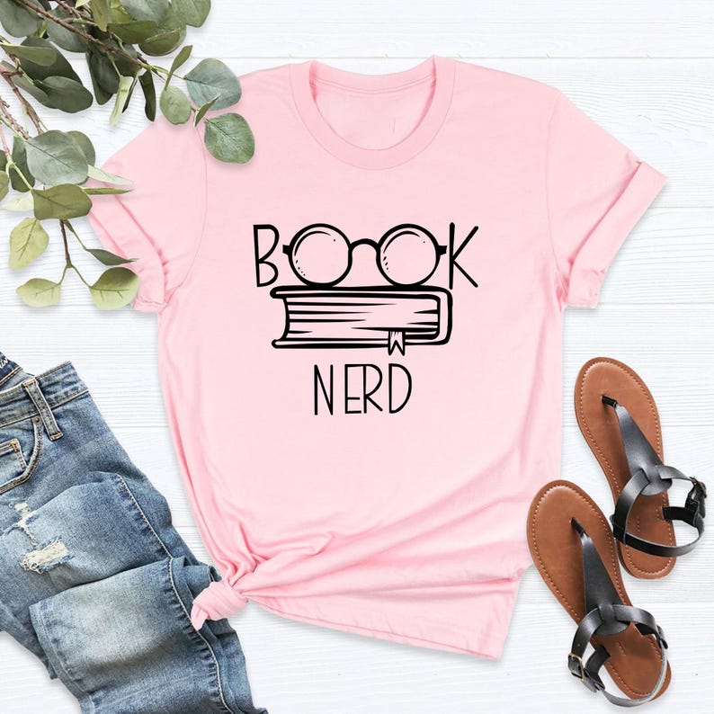 May include: A light pink t-shirt with the words "BOOK NERD" in black. The design features a book with glasses on top. The shirt is paired with blue jeans and brown sandals.