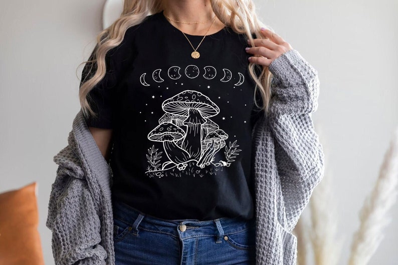 May include: Black t-shirt with a white graphic of mushrooms and a crescent moon phase design. The text "Shroom Queens" is printed on the shirt.