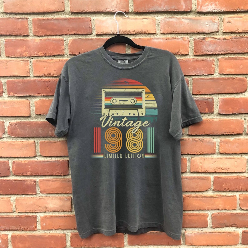 May include: Dark gray t-shirt with a retro design featuring a cassette tape, a sunset, and the text "Vintage 1981 Limited Edition" in a vintage font. The shirt is hanging on a black hanger against a brick wall.