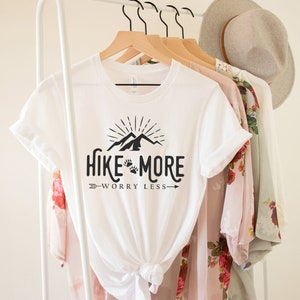 May include: White t-shirt with the text "Hike More Worry Less" in black, featuring a mountain and paw print design. The shirt is hanging on a wooden hanger with other clothing items, including a floral patterned top and a gray hat.