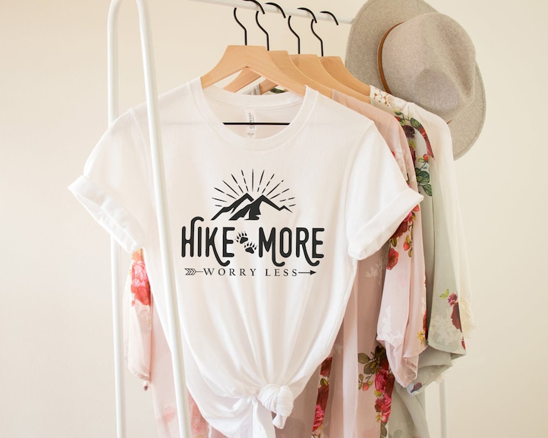 May include: White t-shirt with the text "Hike More Worry Less" in black, featuring a mountain and paw print design. The shirt is hanging on a wooden hanger with other clothing items, including a floral patterned top and a gray hat.