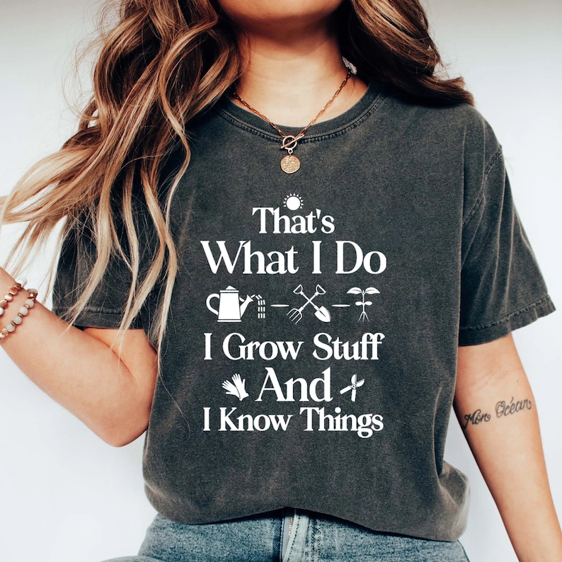 May include: A dark gray t-shirt with white text that reads "That's What I Do I Grow Stuff And I Know Things". The text is surrounded by gardening-themed icons, including a watering can, a shovel, a rake, and a plant.