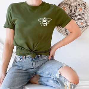 May include: An olive green short-sleeved t-shirt featuring a white line art graphic of a bee on the upper left chest. The shirt has a crew neckline and is styled with a knot at the waist, paired with light blue distressed denim jeans.