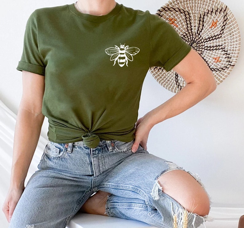 May include: An olive green short-sleeved t-shirt featuring a white line art graphic of a bee on the upper left chest. The shirt has a crew neckline and is styled with a knot at the waist, paired with light blue distressed denim jeans.