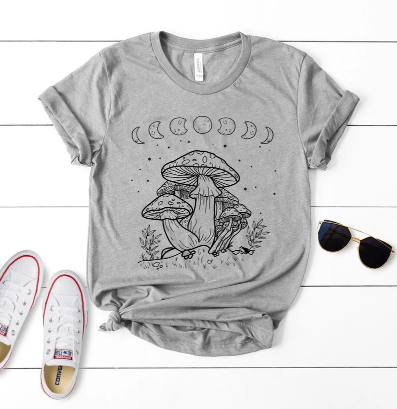 May include: A gray t-shirt with a black and white illustration of mushrooms and a crescent moon design. The shirt has a crew neck and short sleeves.