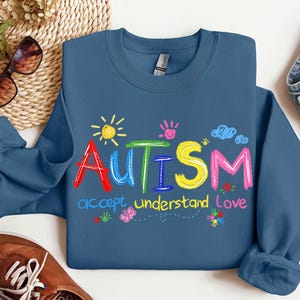 May include: Indigo blue sweatshirt with the word "AUTISM" in colorful, playful letters. Below the word are the words "accept, understand, love". The design includes a sun, handprints, clouds, and flowers.