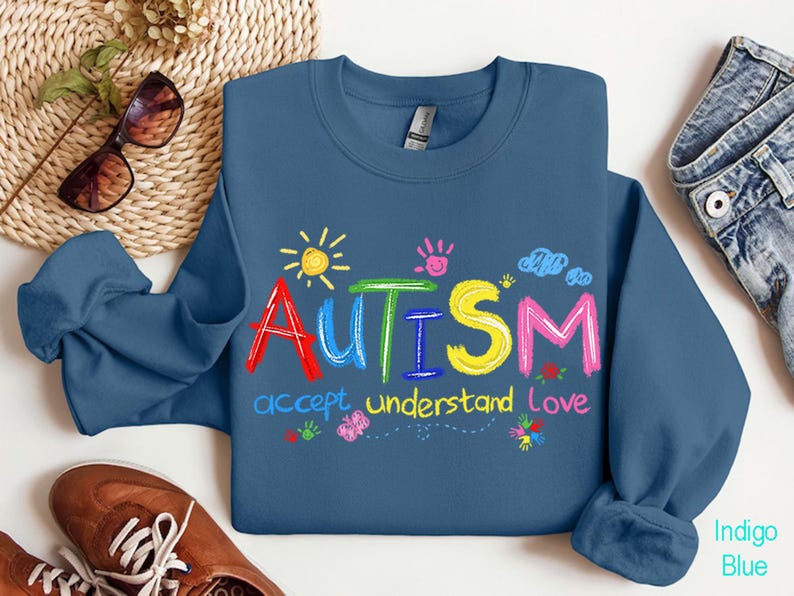 May include: Indigo blue sweatshirt with the word "AUTISM" in colorful, playful letters. Below the word are the words "accept, understand, love". The design includes a sun, handprints, clouds, and flowers.