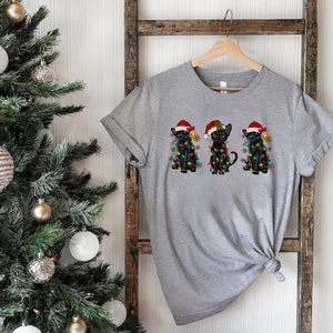 Pode incluir: Uma camiseta cinza com um design de tr&ecirc;s gatos pretos usando chap&eacute;us de Papai Noel e envoltos em luzes de Natal.