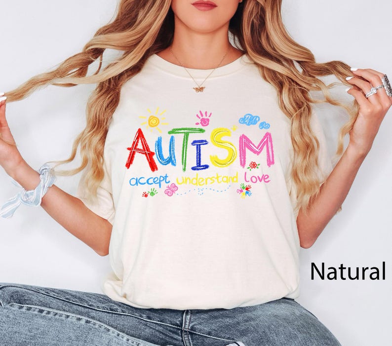 May include: Cream-colored t-shirt with the word "AUTISM" in colorful, hand-drawn letters. Below the word are the words "accept understand love" in a matching style. The design includes a sun, handprint, clouds, and flowers.
