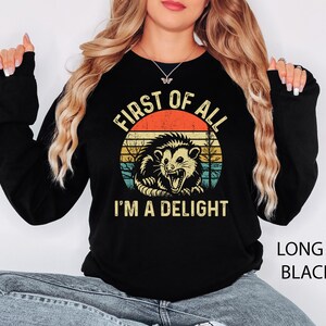 May include: Black long-sleeve sweatshirt featuring a graphic of an opossum with the text "FIRST OF ALL I'M A DELIGHT" in a retro design. The sweatshirt is black with the words "LONG S. BLACK" printed on the bottom right.