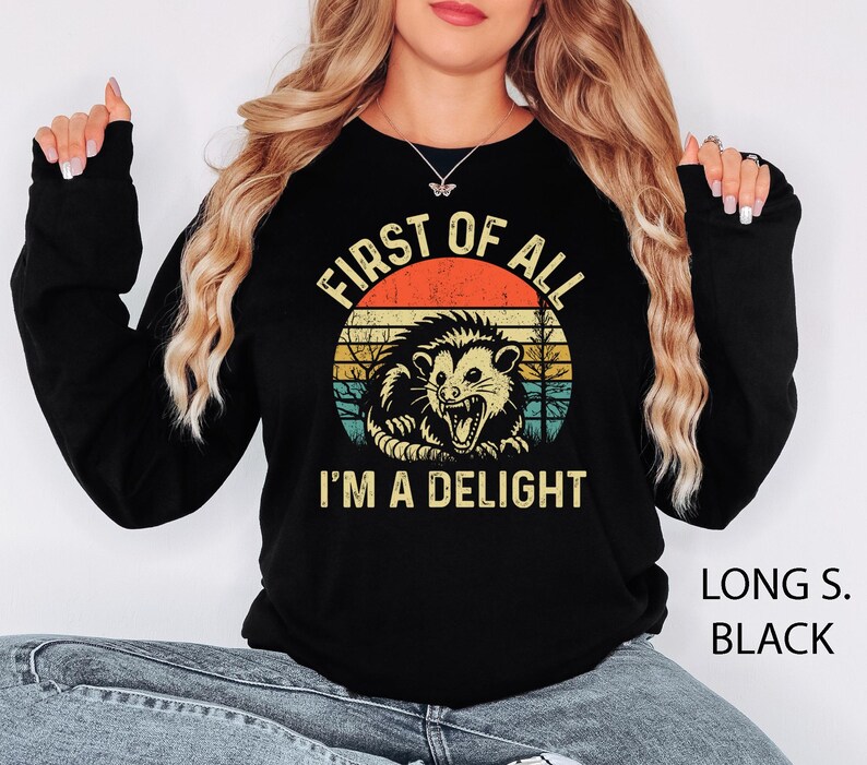 May include: Black long-sleeve sweatshirt featuring a graphic of an opossum with the text "FIRST OF ALL I'M A DELIGHT" in a retro design. The sweatshirt is black with the words "LONG S. BLACK" printed on the bottom right.